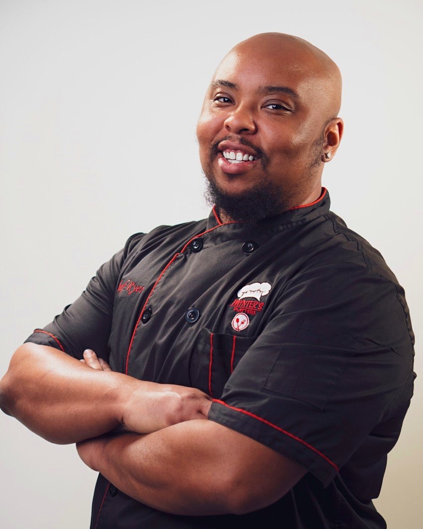 Chef Ryen, founder of Paynter's Platters, smiling in his black and red chef jacket