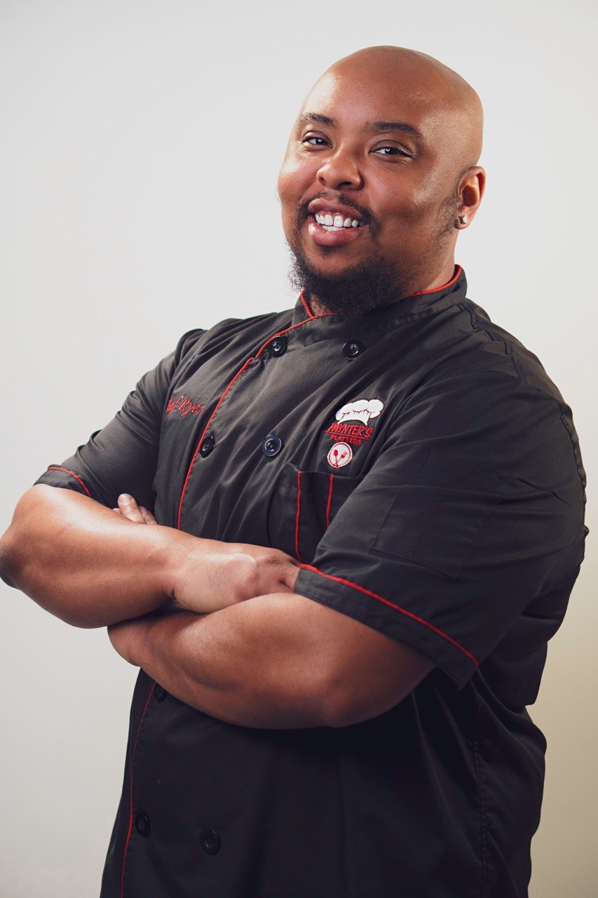 Chef Ryen of Paynter's Platters, arms crossed in his black chef jacket with red piping